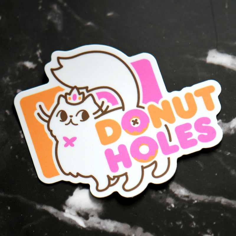 A die-cut sticker featuring a cute white fluffy cat wearing a small pink tiara and bow, with the words “DONUT HOLES” in orange and pink bubble letters over an orange and pink background, placed on a black marble surface.